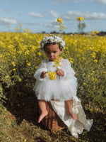 A toddler sits, wearing our Eva flower girl dress, on a basket in a flower field.