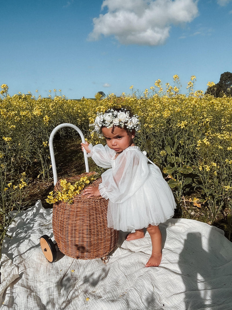 Eva baby flower girl dress with full length tulle sleeves and ivory tulle, is worn by a baby girl. She stands in a yellow flower field and wears our Anais flower crown.