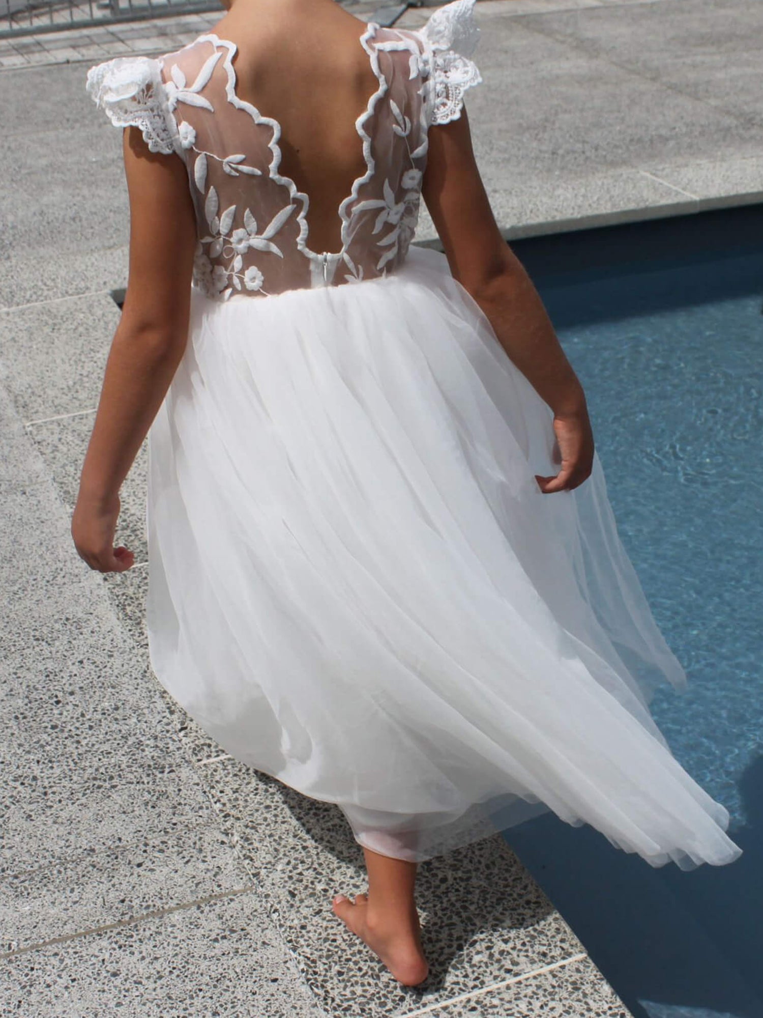 Abigail full length white flower girl dress worn by a young girl who stands beside a pool. Showing the full length soft tulle skirt.