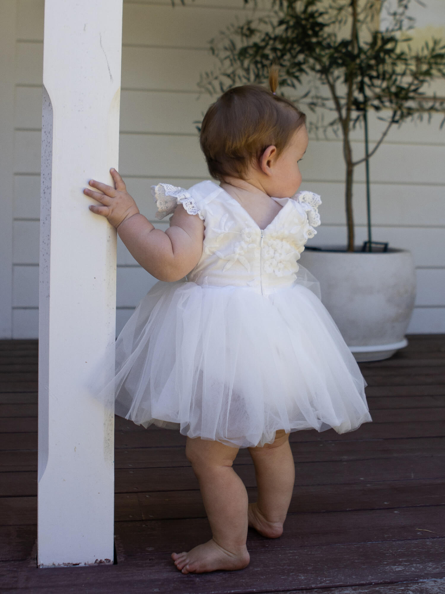 A baby girl stands wearing our Abigail baby flower girl romper in ivory lace and tulle.
