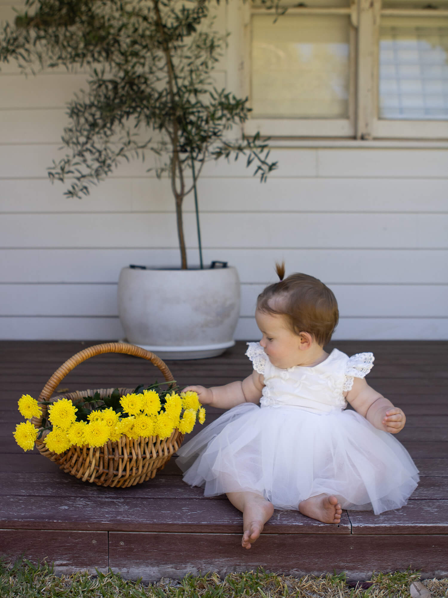 Baby in a white flower girl dress sitting on a wooden bench with a basket of yellow flowers.