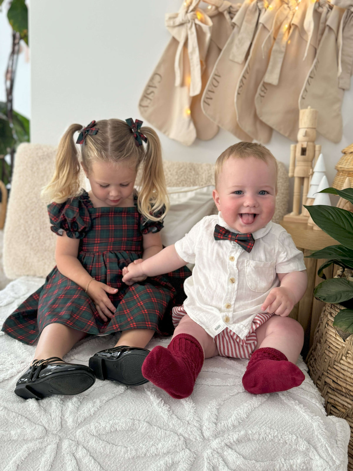 A brother and sister sit wearing matching items from our Christmas collection. Our Mila girls Christmas plaid dress & Christmas plaid boys bow tie.