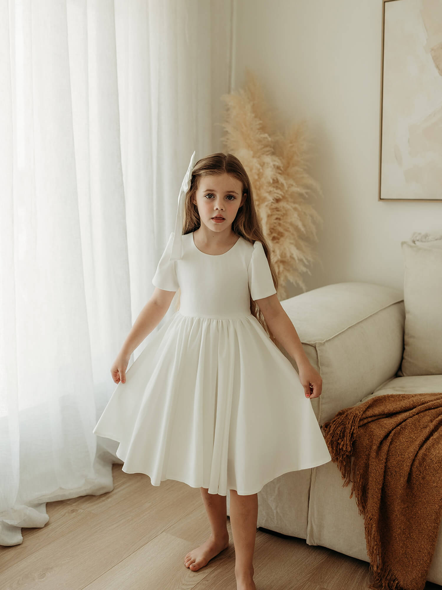 Young girl in a white flower girl dress standing in a room with light-coloured walls and decor.