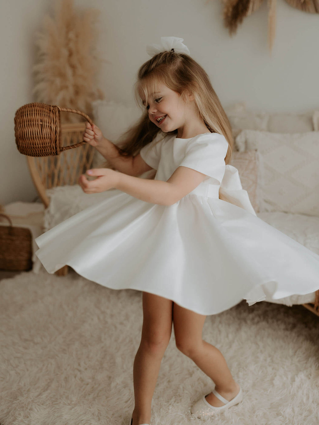 A young girl sits holding a basket. She wears our Charlotte satin flower girl dress and ivory tulle bow in her hair.