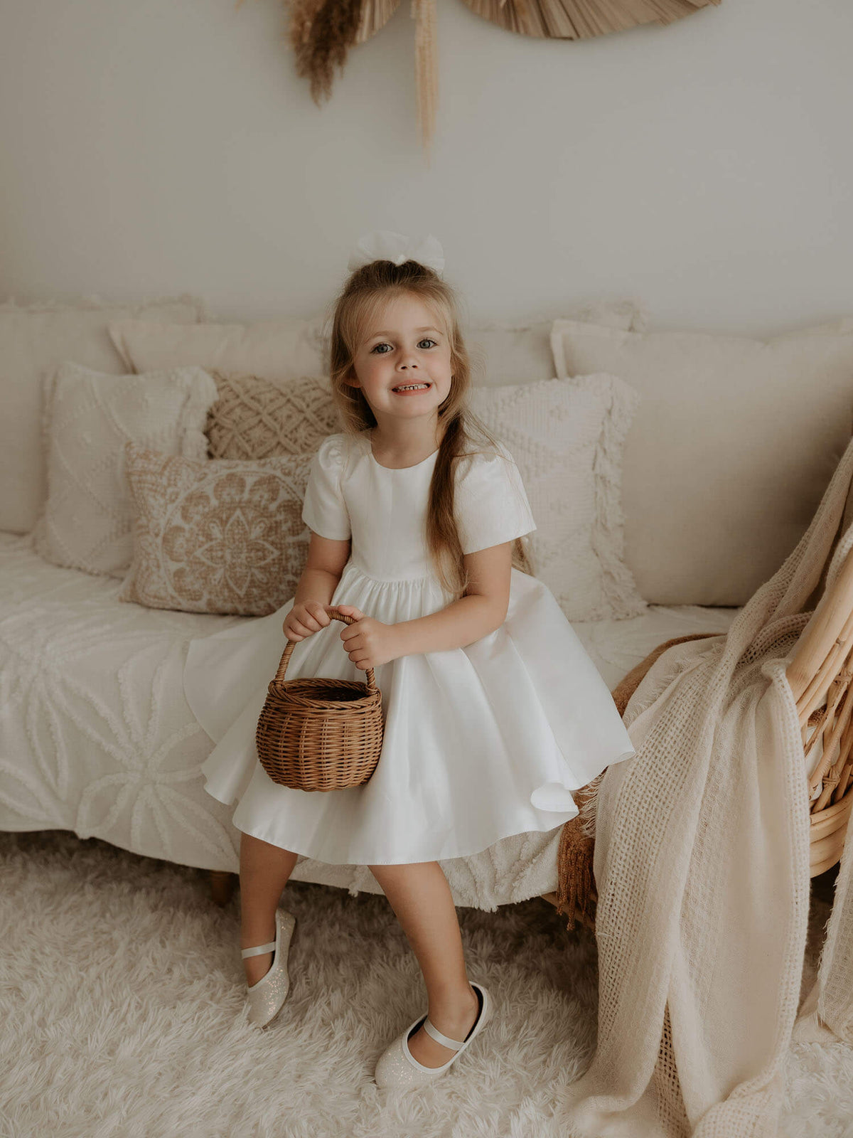 A little girl sits wearing our Charlotte satin dress, perfect for flower girls and communions. She holds a basket and also wears a tulle bow in her hair.