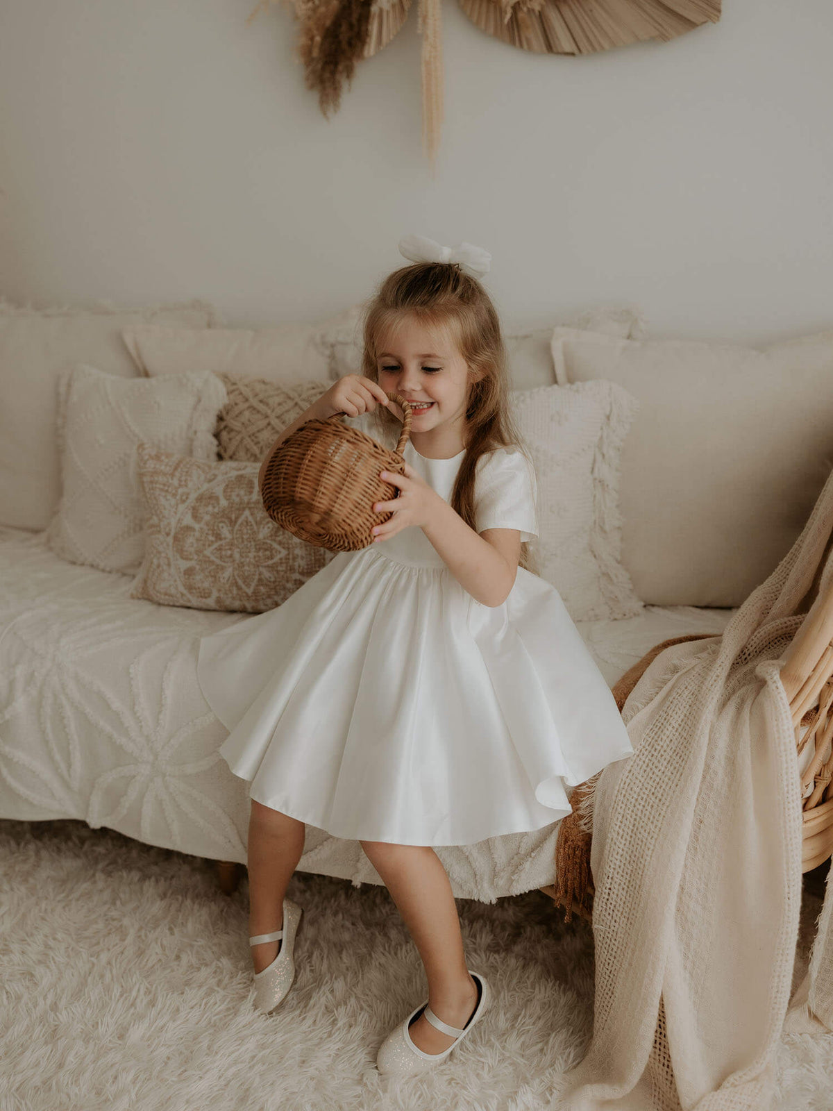 A young girl sits looking into a basket. She wears our Charlotte satin flower girl dress and ivory tulle bow hair clip.