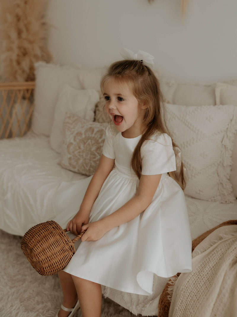 A young girl sits wearing our satin Charlotte dress and ivory tulle bow hair clip.