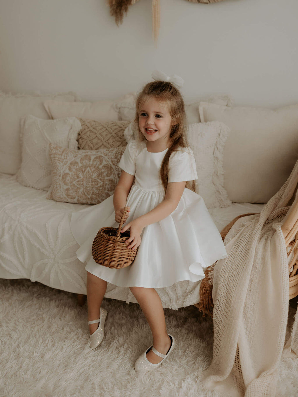 A young girl sits holding a basket. She wears our Charlotte satin flower girl dress and ivory tulle bow in her hair.