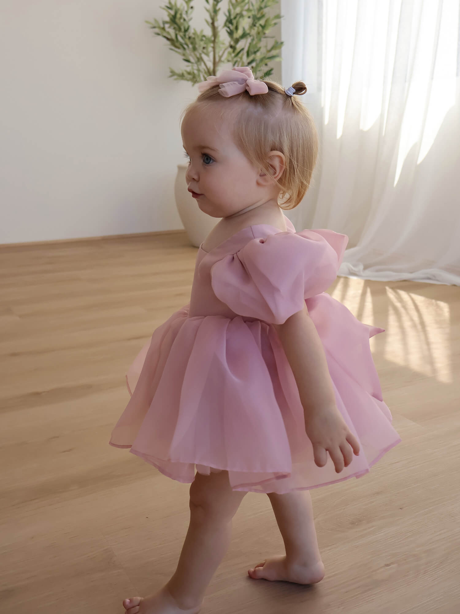 A baby girl wears our blush Cleo baby romper with puff sleeves, along with tulle bows in her hair.