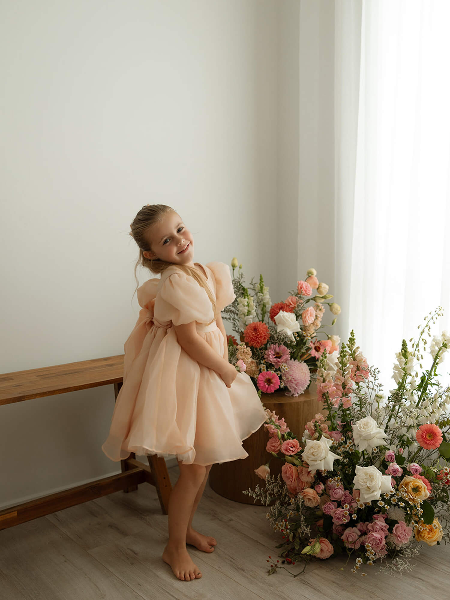 A little girl smiles standing beside colourful flowers. She wears our Cleo champagne puff sleeve flower girl dress.