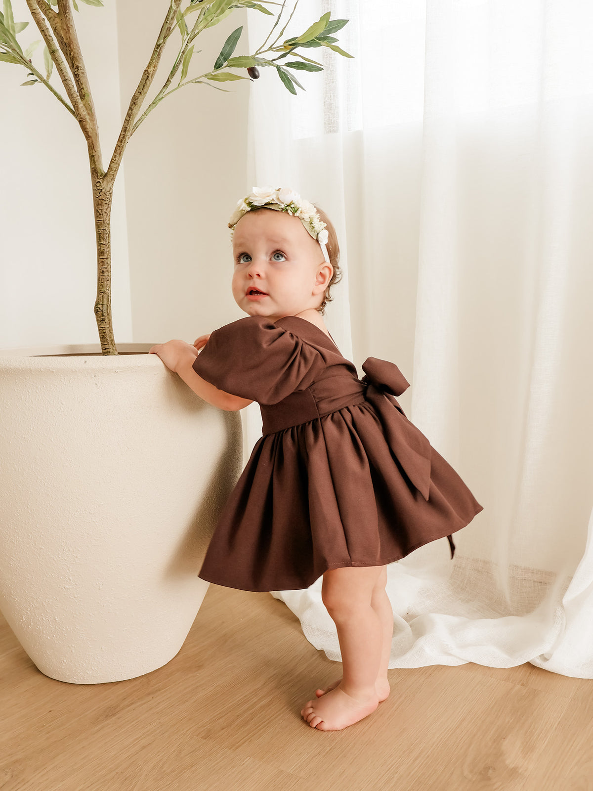 A toddler girl wearing a brown puff-sleeve romper with a bow at the back and a matching flower crown, standing next to a large potted plant in a bright room with white curtains.