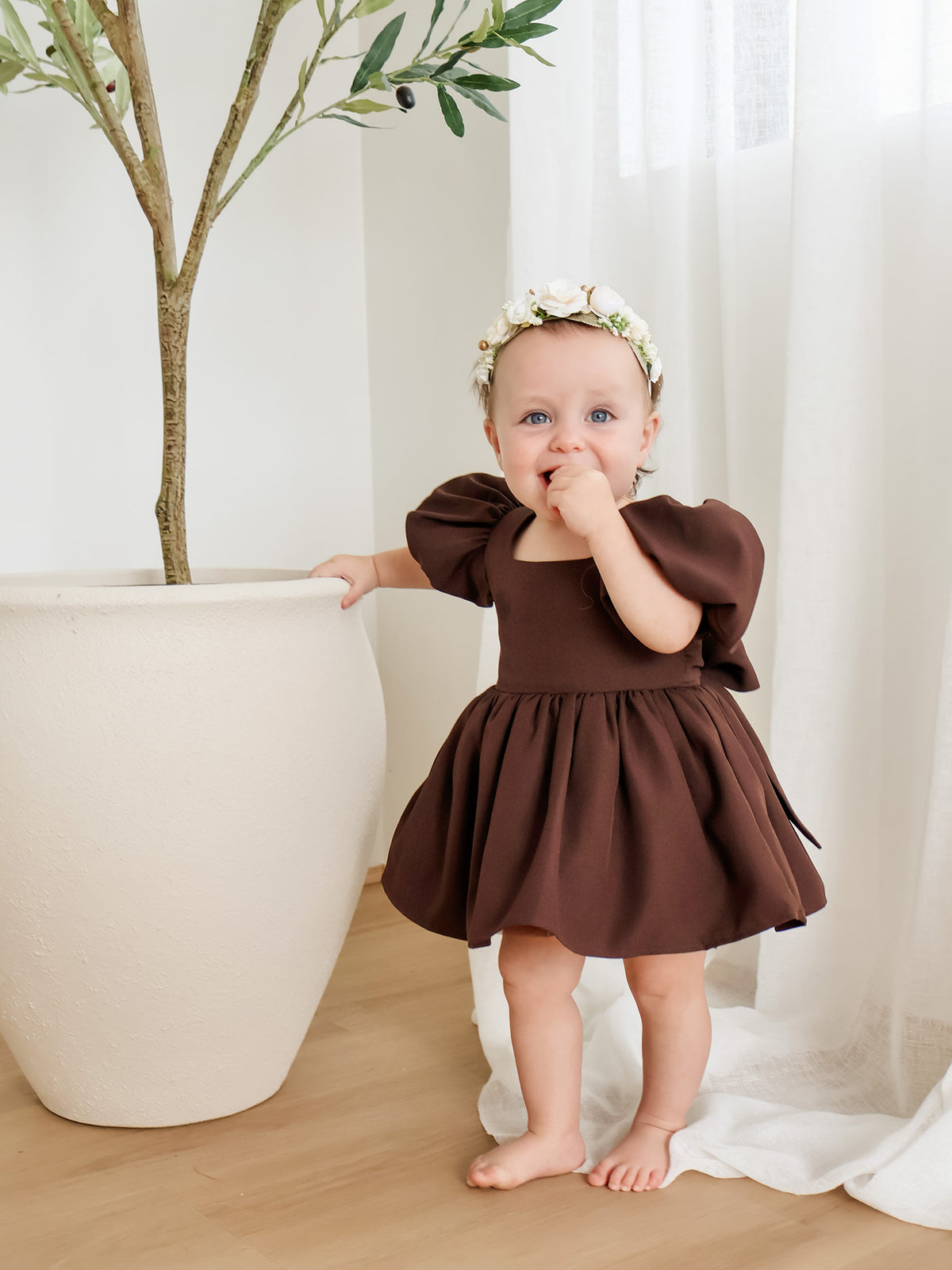 A baby girl wearing a brown puff-sleeve dress and a white floral headband stands in a bright room next to a large white planter.
