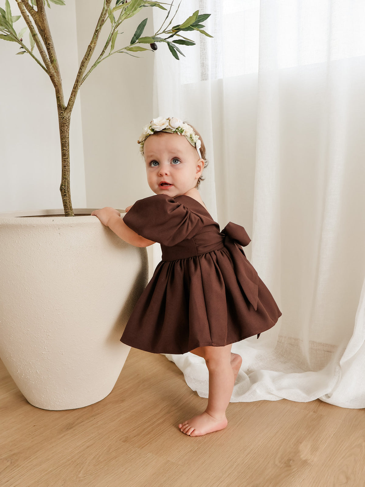A toddle wears our Cleo flower girl romper in chocolate brown crepe. She also wears an ivory floral headband for babies.