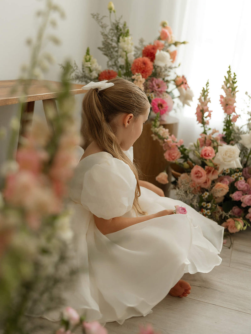 Cream puff sleeved flower girl dress, our Cleo dress in cream organza, is worn by a little girl who is sitting amongst vases of flowers.