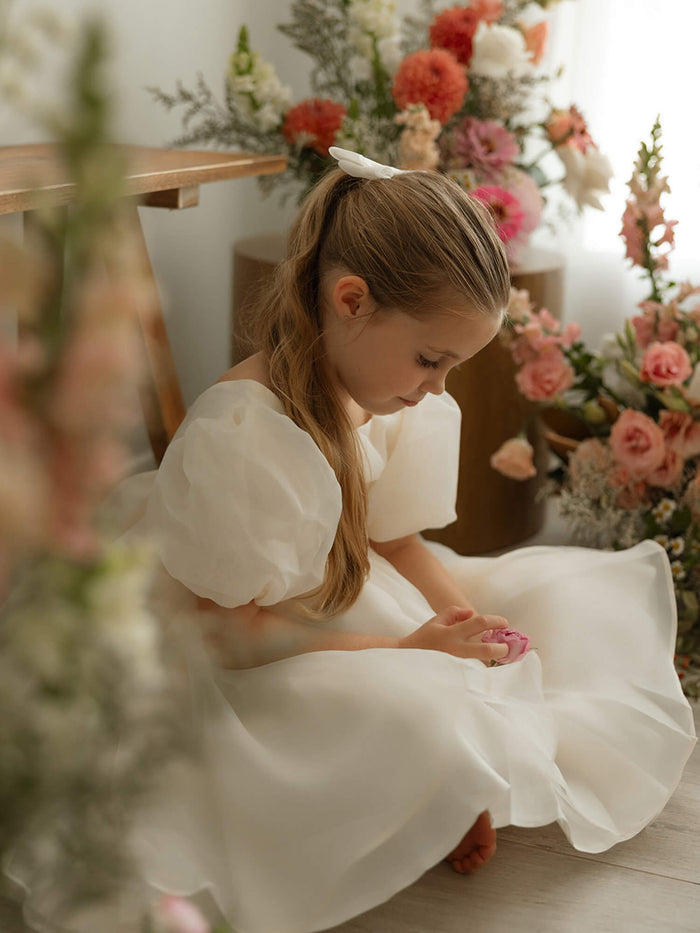 A young girl sits wearing our Cleo cream organza flower girl dress and matching cream organza bow in her hair.