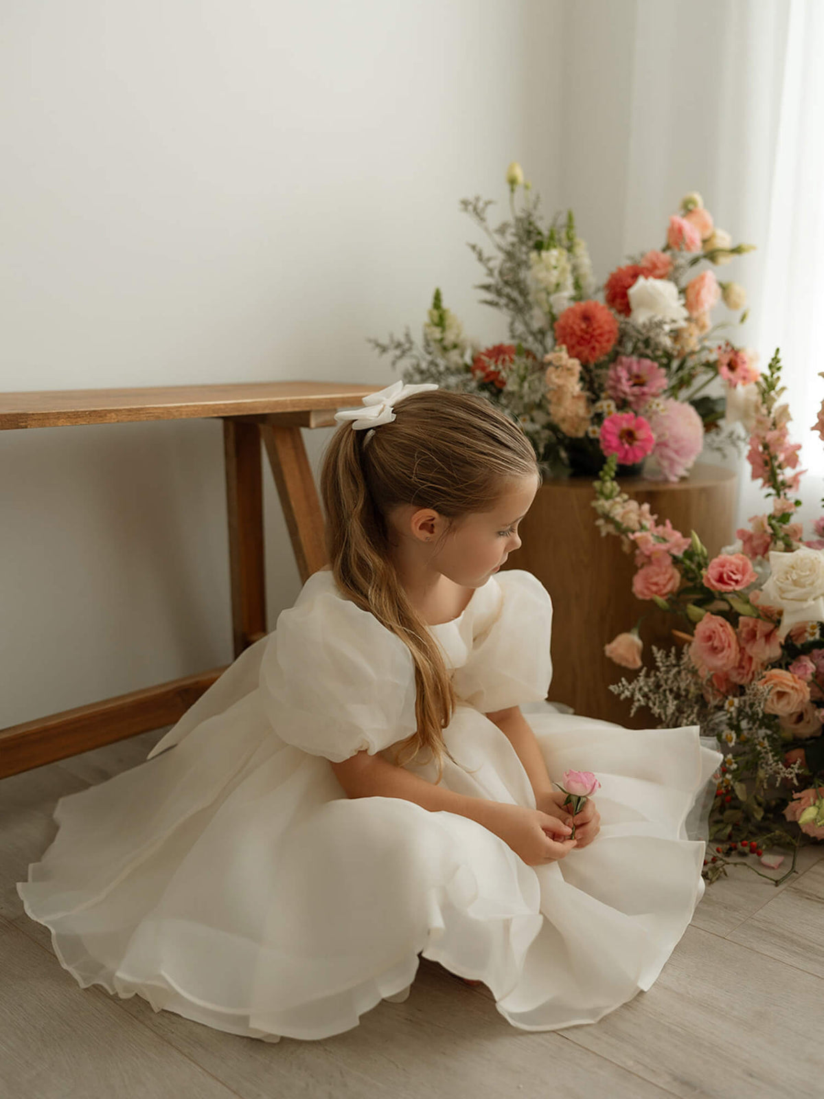 Young flower girl dressed in a formal Cleo gown, ideal for elegant wedding ceremonies. She sits near vases of flowers.