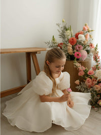 Cleo cream organza flower girl dress is worn by a young girl, who sits on the floor beside vases of flowers.