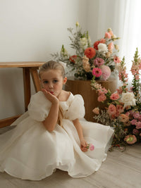 A little girl wearing the Cleo flower girl dress in puff-sleeved cream organza.