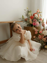 A cream puff sleeve flower girl dress in organza, styled for a timeless wedding look, is worn by a young girl.