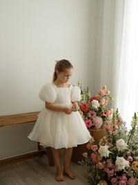 A little girl stands beside vases of flowers wearing our cream Cleo flower girl dress with puff sleeves.