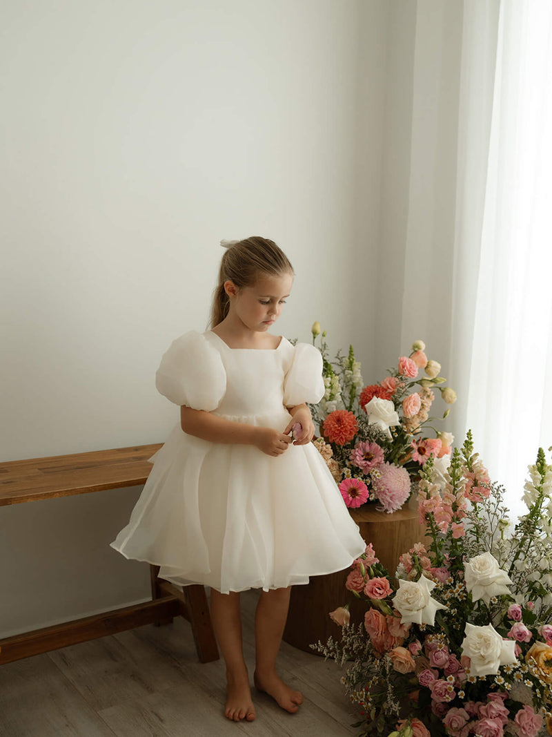 A little girl stands beside vases of flowers wearing our cream Cleo flower girl dress with puff sleeves.