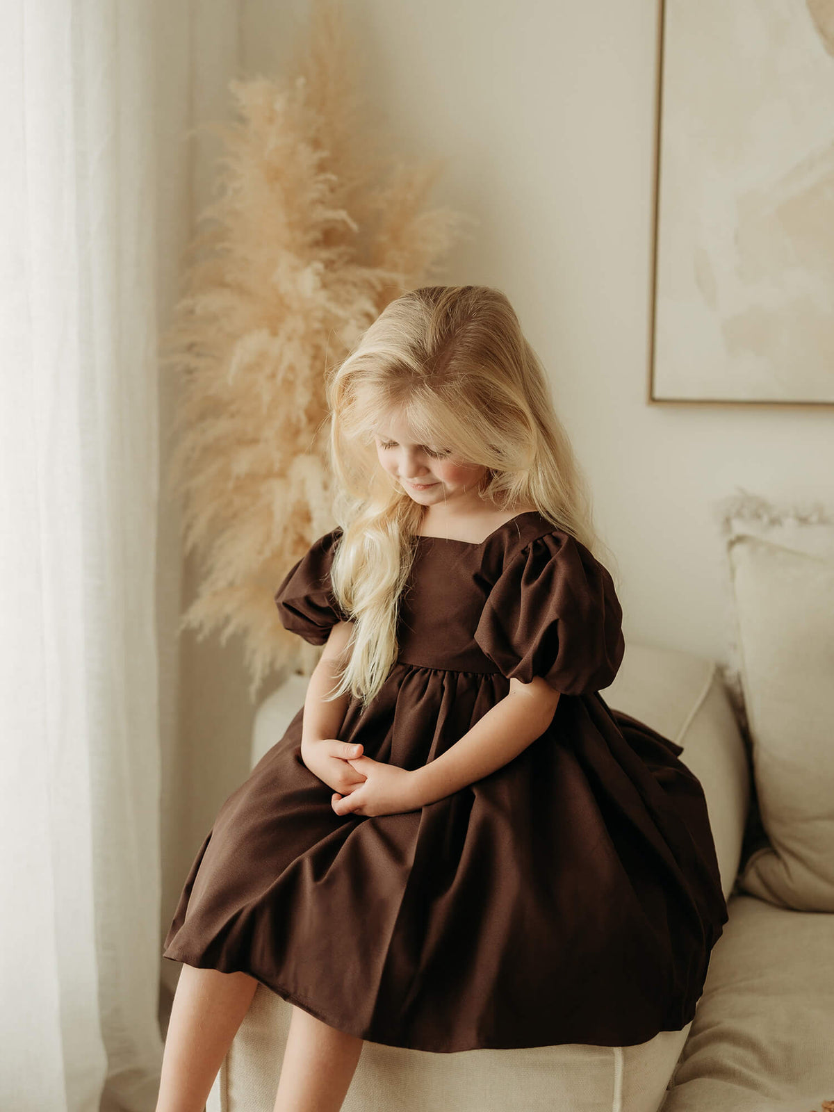 A young girl wearing a dark brown puff-sleeve flower girl dress sits on a beige sofa in a softly lit room with neutral decor.