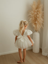 A little girl wears our Cleo baby flower girl romper, showing the puff sleeves and bow at the back.
