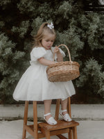 Cleo linen flower girl dress is worn by a young girl. She sits on a stool holding a flower girl basket, with a linen bow in her hair.