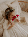 A young girl holds flowers, wearing our Linen puff sleeve Cleo flower girl dress and Blythe floral crown.