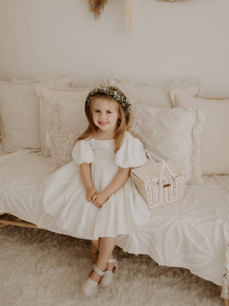 A young girl sits smiling, wearing our Cleo puff sleeve flower girl dress in linen.