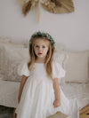 Cleo linen puff sleeve flower girl dress is worn by a young girl. she holds a flower girl basket and wears a floral wreath in her hair.