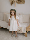 Cleo linen flower girl dress with puff sleeves is worn by a young girl. She holds a basket and wears a floral wreath.