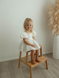 A little girl sits on a stool wearing our ivory linen Cleo flower girl romper.