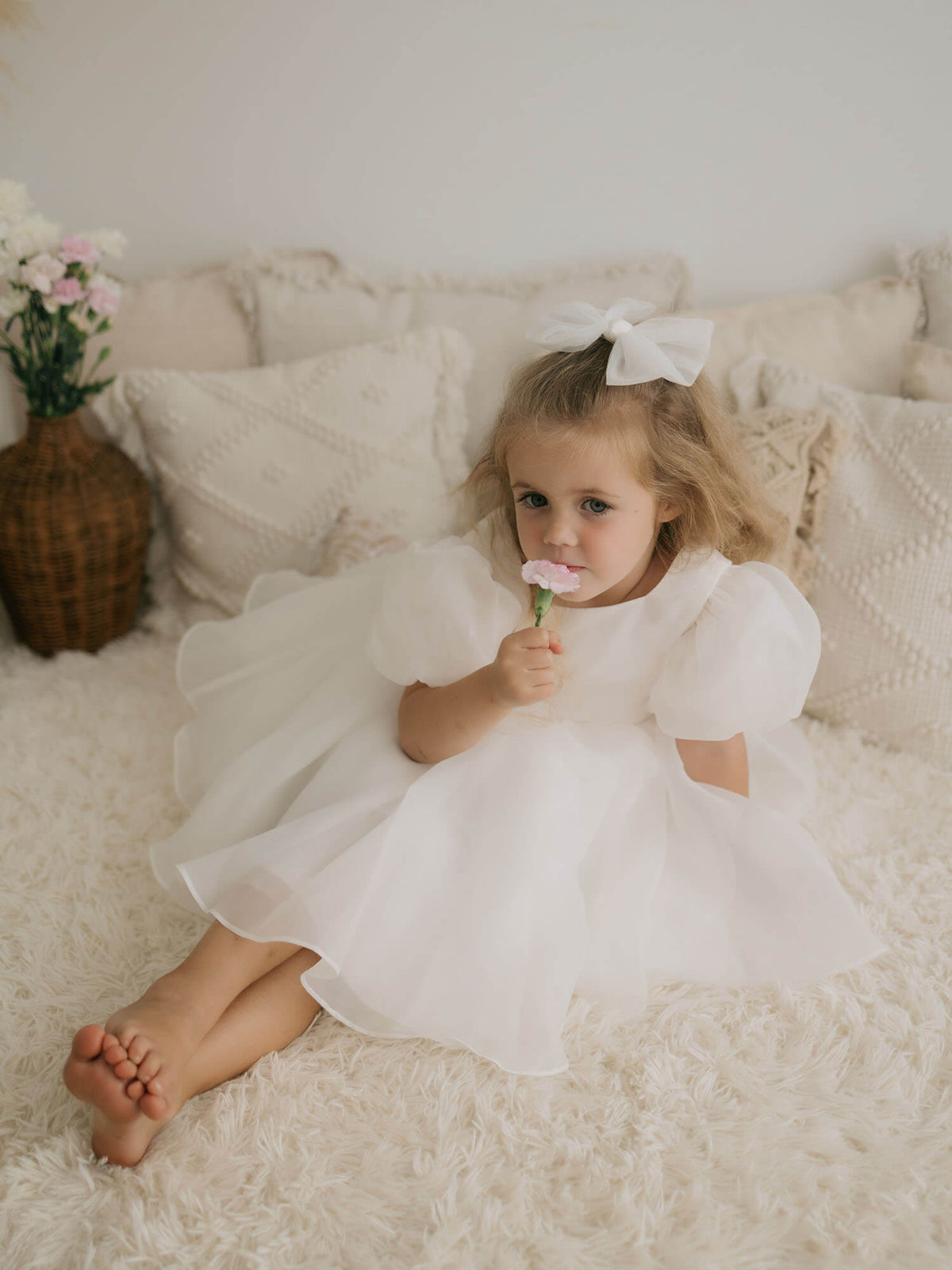 A girl sits smelling a flower. She wears our Cleo puff sleeve flower girl dress and medium ivory tulle bow.