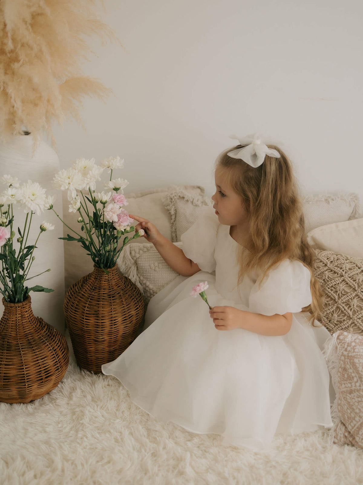 Cleo ivory organza flower girl dress is worn by a young girl who sits playing with flowers. She also wears our medium ivory tulle bow hairclip in her hair.