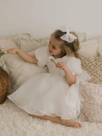 A girl sits wearing our organza Cleo flower girl dress and medium ivory tulle bow hair clip.