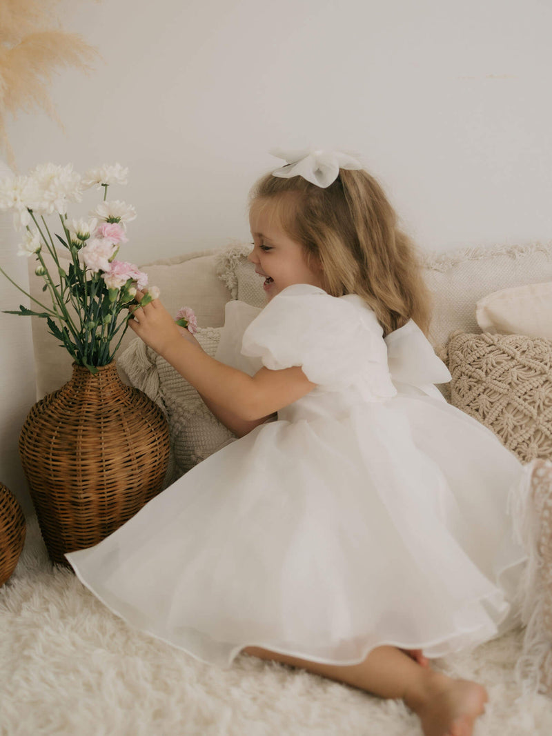 A little girl laughs, playing with flowers. She wears our organza puff sleeve Cleo flower girl dress with organza bow at the back.