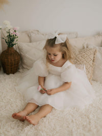 A young girl sits holding a flower. She wears our Cleo organza flower girl dress and ivory medium tulle bow, in her hair.