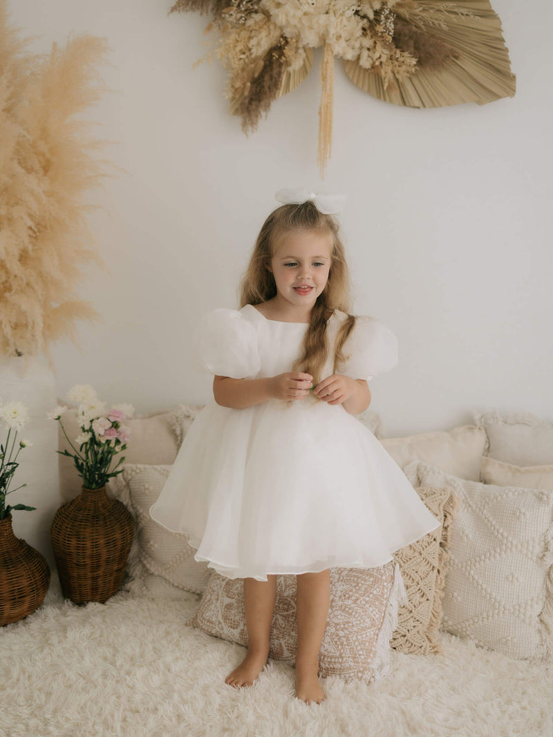 A young flower girl wearing our Cleo white puff sleeve flower girl dress sits with her sister who wears our Emma blush romper.