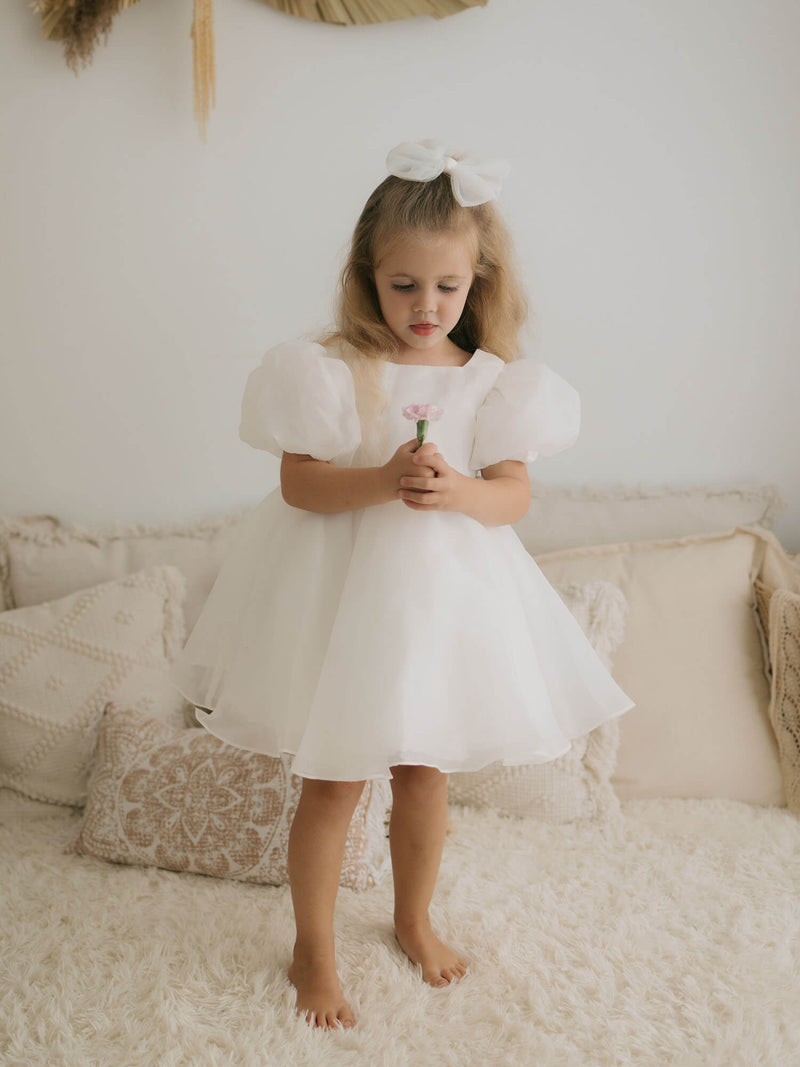 A young flower girl sits in our Cleo ivory puff sleeve flower girl dress. She also wears a tulle bow in her hair.