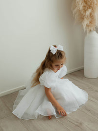 A young girl sits wearing our Cleo pure white flower girl dress and organza hair bow in pure white.