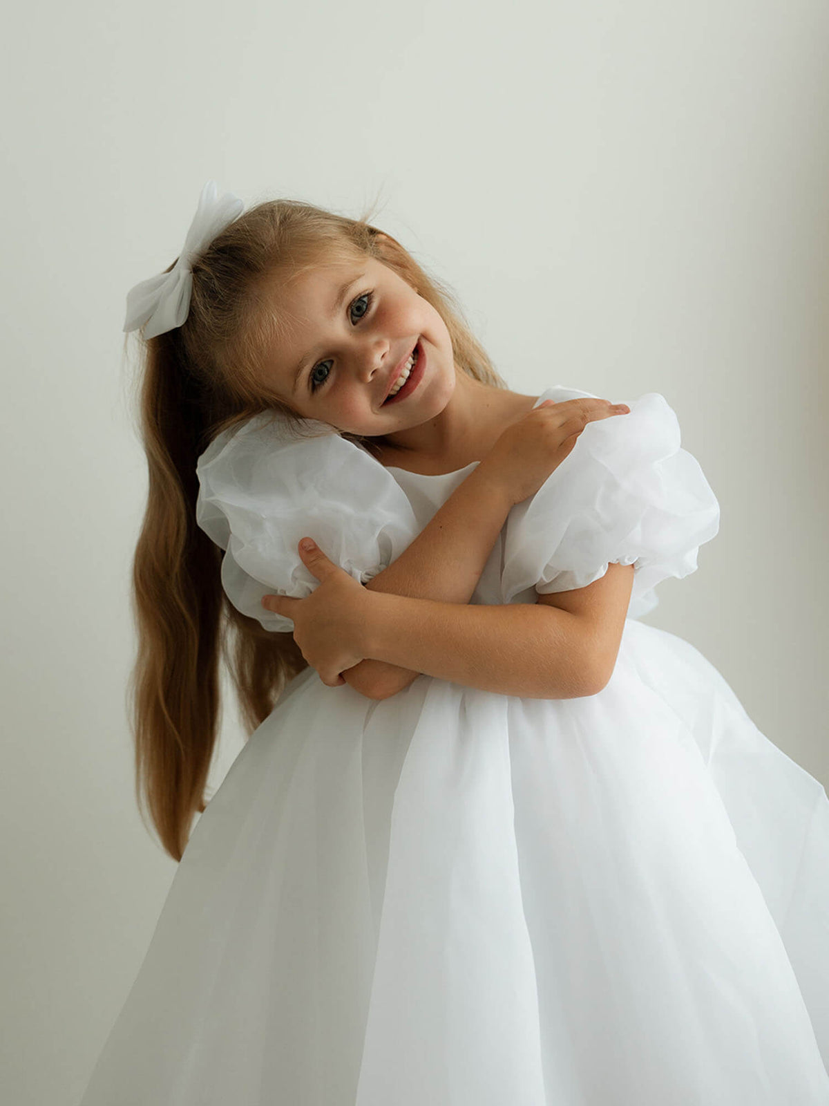 A young girl wears our Cleo pure white flower girl dress and matching organza bow.