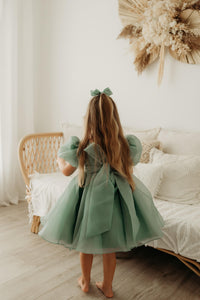 The back of our Cleo flower girl dress in jade, worn by a young girl. Showing the oversized bow at the back and the puff sleeves.