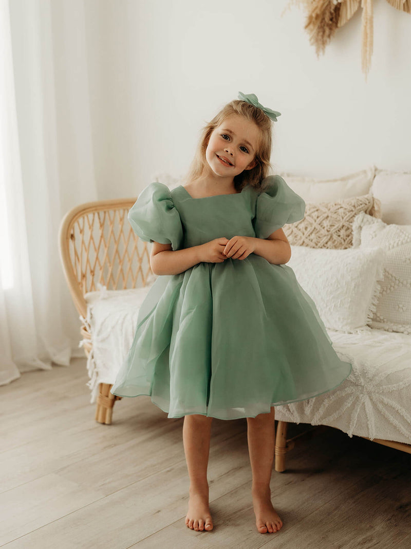 Cleo jade green flower girl dress with puff sleeves is worn by a girl. She also wears a matching organza bow in her hair.