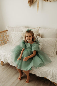 Cleo sage green puff sleeve flower girl dress is worn by a young girl who is sitting on a couch.