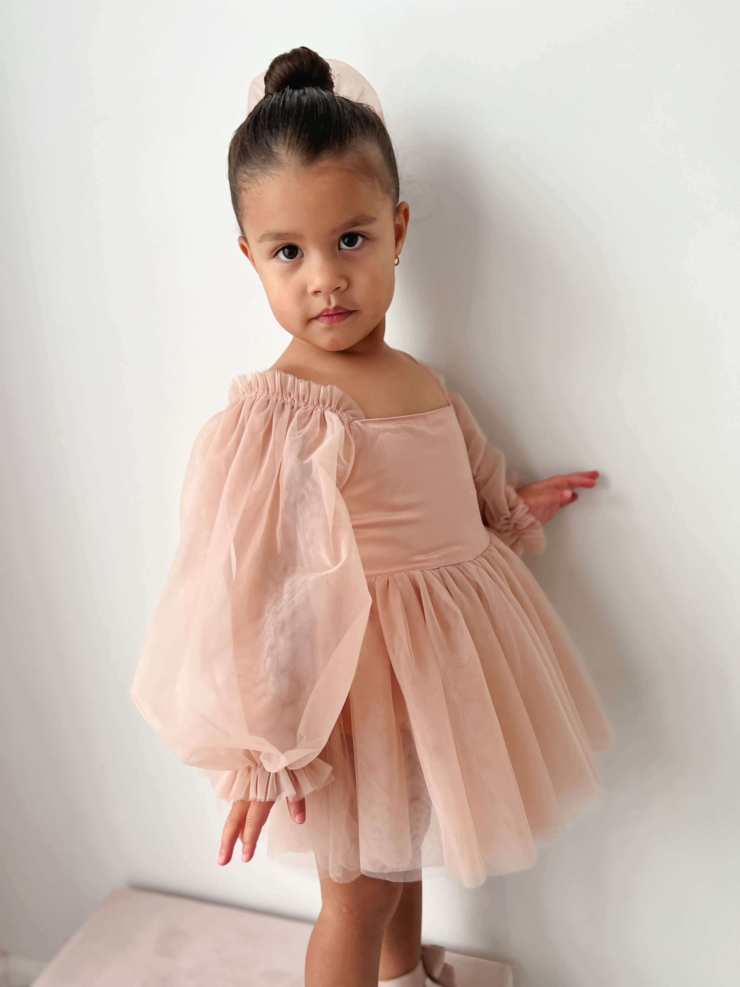 A little girl stands wearing our Eva champagne baby flower girl dress and matching tulle hair bow.