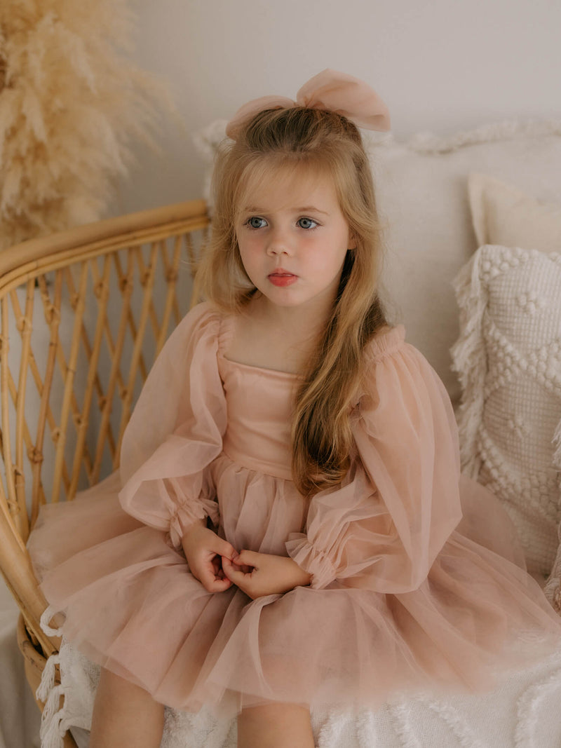Eva tulle sleeve flower girl dress in champagne is worn by a young girl, along with a large tulle bow hair clip in champagne.