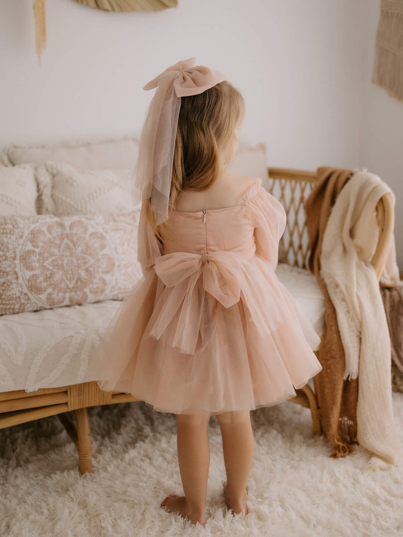 Eva flower girl dress in champagne is worn by a young girl along with a large tulle bow hair clip in champagne.
