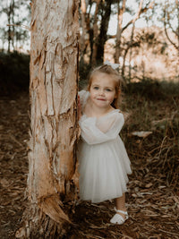 A flower girl peeks around from the side of a tree wearing our Eva flower girl dress with full length tulle sleeves.