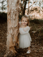 A flower girl peeks around from the side of a tree wearing our Eva flower girl dress with full length tulle sleeves.